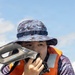 A person wearing an orange life vest and digital camouflage boonie hat holds an Aquapac waterproof case or device up to their face. The individual appears to be on or near a watercraft, suggesting a maritime or water-based activity. The image is a close-up portrait taken outdoors under a partly cloudy sky.