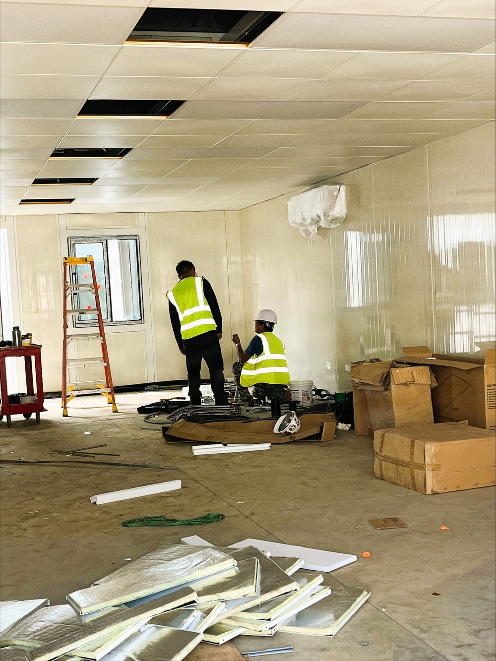 Two construction workers wearing high-visibility yellow vests are working inside a prefabricated modular building or portable office unit under active fit-out. The interior shows white panel walls and a drop ceiling with several open tile slots, indicating ongoing electrical or HVAC installation work. The space is cluttered with cardboard boxes, insulated ceiling panels, tools, and a stepladder, consistent with an early-stage interior fit-out.