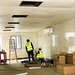 Two construction workers wearing high-visibility yellow vests are working inside a prefabricated modular building or portable office unit under active fit-out. The interior shows white panel walls and a drop ceiling with several open tile slots, indicating ongoing electrical or HVAC installation work. The space is cluttered with cardboard boxes, insulated ceiling panels, tools, and a stepladder, consistent with an early-stage interior fit-out.