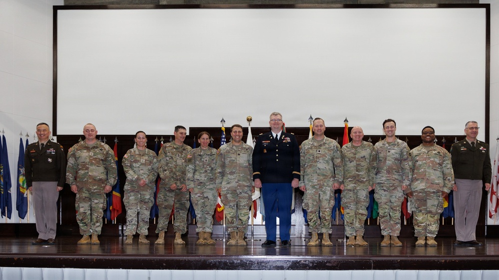 A formal military group photograph taken on a stage with multiple state and unit flags displayed in the background. Approximately 13 military personnel are posed in a single row, with one individual in the center wearing dress blue trousers distinguishing them from the others in standard Army Combat Uniforms (ACU/OCP). The setting appears to be an official ceremony or formal event held indoors.