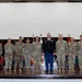 A formal military group photograph taken on a stage with multiple state and unit flags displayed in the background. Approximately 13 military personnel are posed in a single row, with one individual in the center wearing dress blue trousers distinguishing them from the others in standard Army Combat Uniforms (ACU/OCP). The setting appears to be an official ceremony or formal event held indoors.