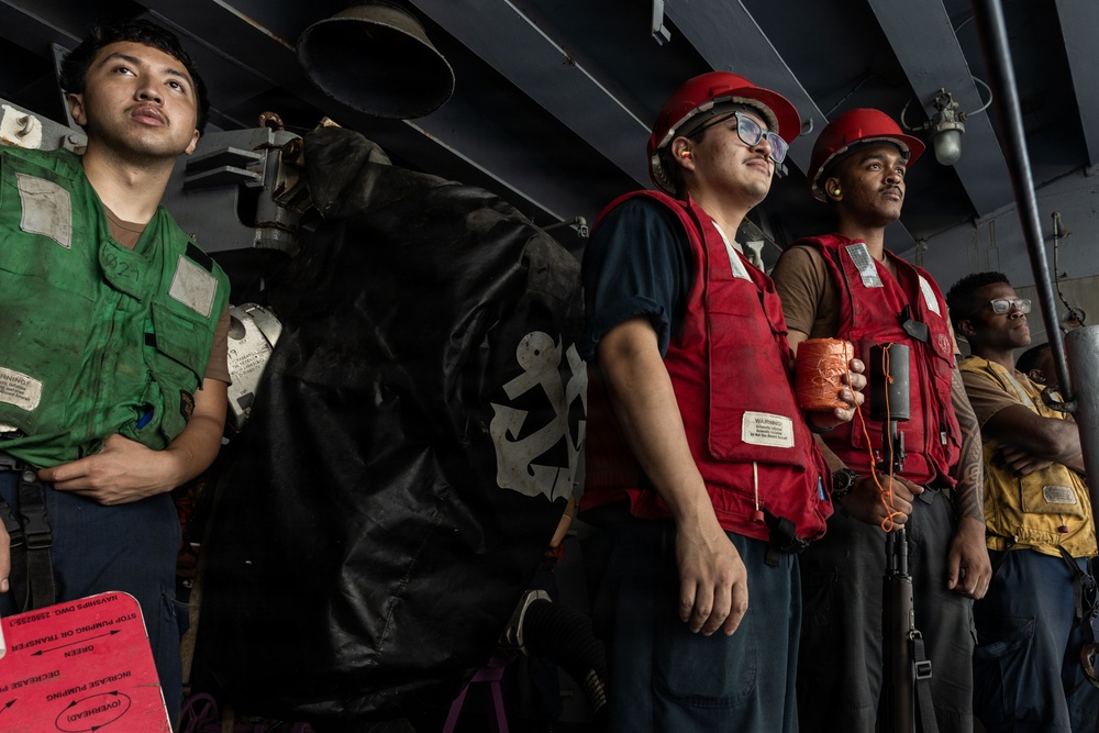 A group of military personnel, likely U.S. Navy sailors, are standing at attention or observing an operation aboard a naval vessel. They are wearing color-coded safety vests and hard hats, indicating assigned roles during a shipboard evolution such as an underway replenishment or deck operation. The setting appears to be an interior deck or hangar bay area of a warship.
