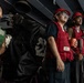 A group of military personnel, likely U.S. Navy sailors, are standing at attention or observing an operation aboard a naval vessel. They are wearing color-coded safety vests and hard hats, indicating assigned roles during a shipboard evolution such as an underway replenishment or deck operation. The setting appears to be an interior deck or hangar bay area of a warship.