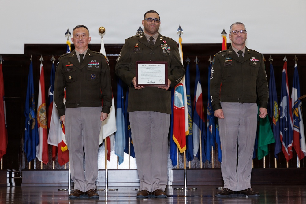 Three U.S. Army soldiers stand on a stage during a formal military ceremony. The central figure holds a framed Certificate of Appreciation, flanked by two higher-ranking officers. Multiple state and organizational flags are displayed in the background.