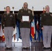 Three U.S. Army soldiers stand on a stage during a formal military ceremony. The central figure holds a framed Certificate of Appreciation, flanked by two higher-ranking officers. Multiple state and organizational flags are displayed in the background.