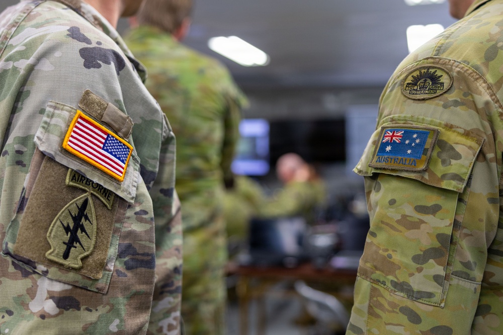 The image shows the upper arms/shoulders of two military personnel in close-up, one wearing a US Army uniform with Airborne and unit patches, and the other wearing an Australian Army uniform with national flag patch. Both soldiers are dressed in MultiCam/OCP camouflage pattern uniforms. The background shows a blurred indoor military operations or briefing room environment with other personnel visible.