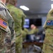 The image shows the upper arms/shoulders of two military personnel in close-up, one wearing a US Army uniform with Airborne and unit patches, and the other wearing an Australian Army uniform with national flag patch. Both soldiers are dressed in MultiCam/OCP camouflage pattern uniforms. The background shows a blurred indoor military operations or briefing room environment with other personnel visible.