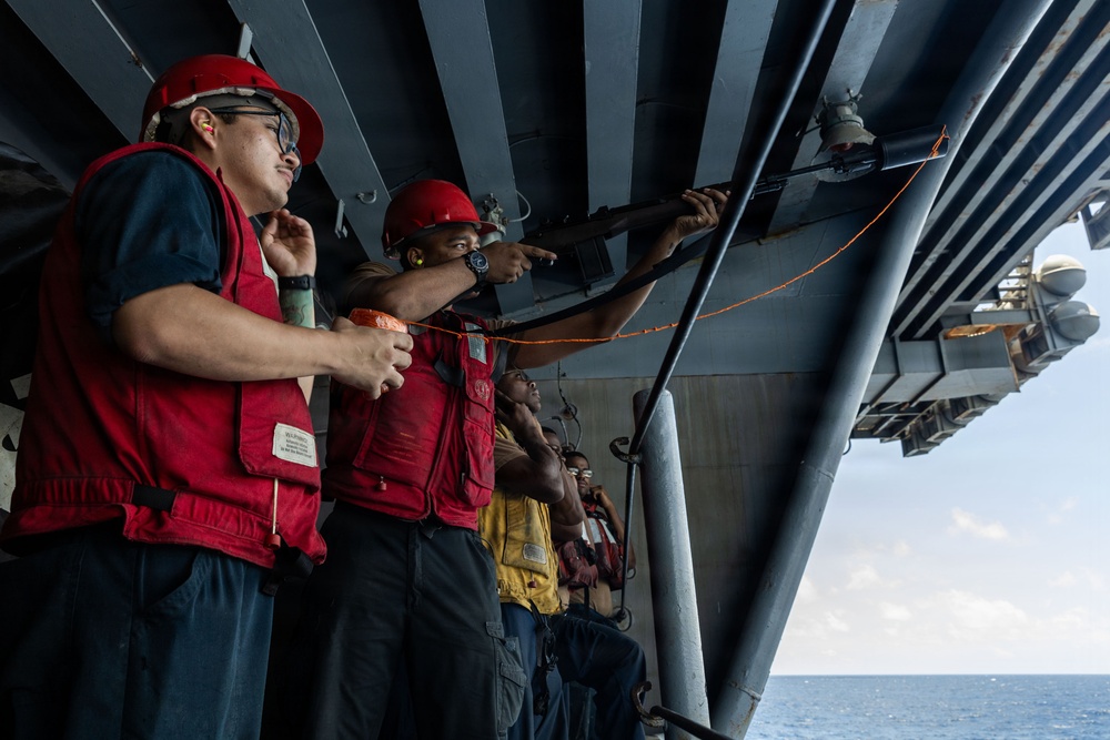 A group of military personnel, likely U.S. Navy sailors, are standing on the exterior deck or catwalk of a large naval vessel at sea. They are wearing red hard hats and red safety vests, with one sailor pointing toward equipment mounted on the ship's superstructure. The setting appears to be an active training or inspection exercise conducted underway.