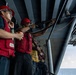 A group of military personnel, likely U.S. Navy sailors, are standing on the exterior deck or catwalk of a large naval vessel at sea. They are wearing red hard hats and red safety vests, with one sailor pointing toward equipment mounted on the ship's superstructure. The setting appears to be an active training or inspection exercise conducted underway.