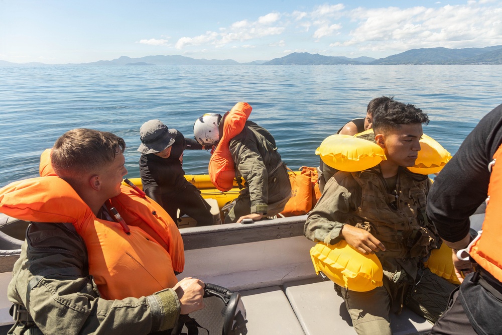 Several individuals in military-style olive/green uniforms are aboard a metal boat on calm open water, with mountainous terrain visible in the background. The personnel are wearing orange and yellow inflatable life vests/flotation devices, suggesting a water survival or rescue training exercise. Some individuals appear to be handling or deploying an inflatable yellow raft.