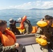 Several individuals in military-style olive/green uniforms are aboard a metal boat on calm open water, with mountainous terrain visible in the background. The personnel are wearing orange and yellow inflatable life vests/flotation devices, suggesting a water survival or rescue training exercise. Some individuals appear to be handling or deploying an inflatable yellow raft.
