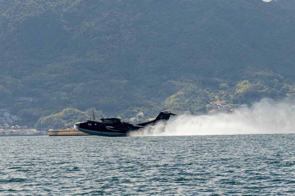 A dark-colored flying boat or seaplane, marked with the number '08', is performing a water takeoff or landing run on a calm bay or inlet. The aircraft is generating a large spray wake as it accelerates across the water surface. A forested hillside with scattered buildings is visible in the background.