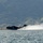 U.S. Marines, JMSDF Sailors reinforce maritime readiness during Rescue Flag 26-1 A dark-colored flying boat or seaplane, marked with the number '08', is performing a water takeoff or landing run on a calm bay or inlet. The aircraft is generating a large spray wake as it accelerates across the water surface. A forested hillside with scattered buildings is visible in the background.