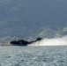 A dark-colored flying boat or seaplane, marked with the number '08', is performing a water takeoff or landing run on a calm bay or inlet. The aircraft is generating a large spray wake as it accelerates across the water surface. A forested hillside with scattered buildings is visible in the background.