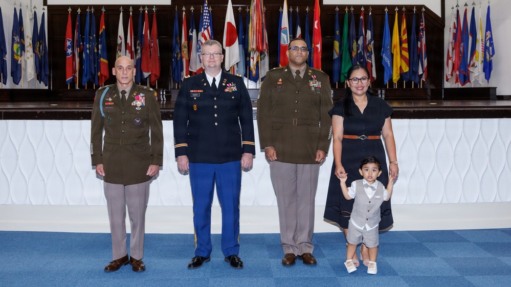 A formal military ceremony photograph featuring three U.S. Army personnel in uniform alongside a woman in civilian attire and a young child. The group is posed in front of a large display of military and state flags, suggesting an official promotion or award ceremony. The setting appears to be a formal military hall or auditorium.
