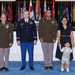 A formal military ceremony photograph featuring three U.S. Army personnel in uniform alongside a woman in civilian attire and a young child. The group is posed in front of a large display of military and state flags, suggesting an official promotion or award ceremony. The setting appears to be a formal military hall or auditorium.