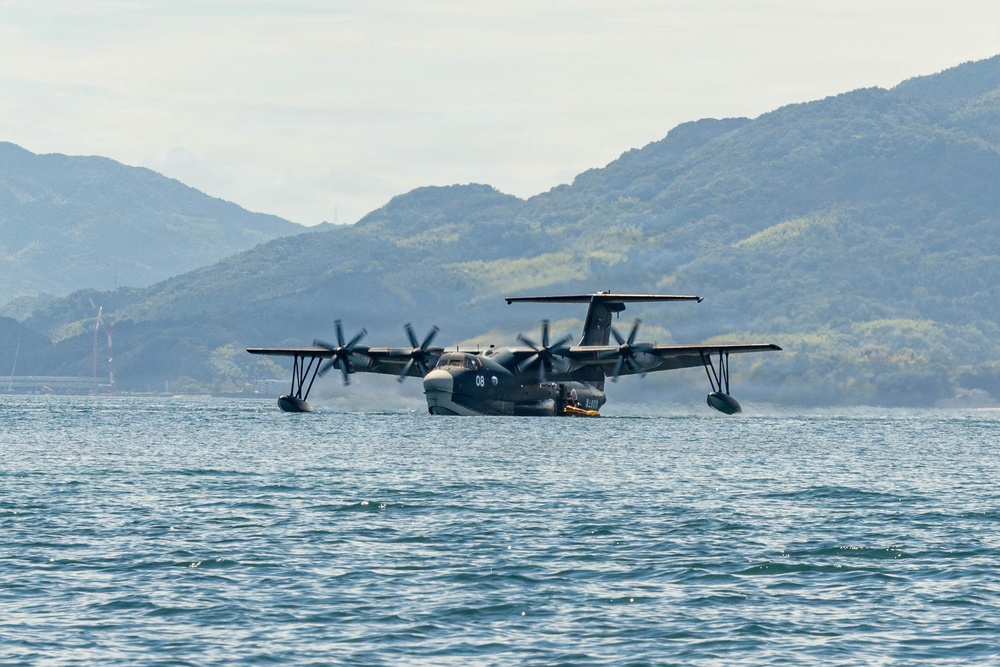 A large military flying boat or amphibious aircraft is taxiing on open water, identifiable by the number '08' on its fuselage. The aircraft features four turboprop engines and wingtip floats, consistent with a Japan Maritime Self-Defense Force US-2 rescue aircraft. It is set against a backdrop of green, forested mountains under a hazy sky.