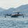 U.S. Marines, JMSDF Sailors reinforce maritime readiness during Rescue Flag 26-1 A large military flying boat or amphibious aircraft is taxiing on open water, identifiable by the number '08' on its fuselage. The aircraft features four turboprop engines and wingtip floats, consistent with a Japan Maritime Self-Defense Force US-2 rescue aircraft. It is set against a backdrop of green, forested mountains under a hazy sky.