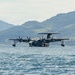 A large military flying boat or amphibious aircraft is taxiing on open water, identifiable by the number '08' on its fuselage. The aircraft features four turboprop engines and wingtip floats, consistent with a Japan Maritime Self-Defense Force US-2 rescue aircraft. It is set against a backdrop of green, forested mountains under a hazy sky.