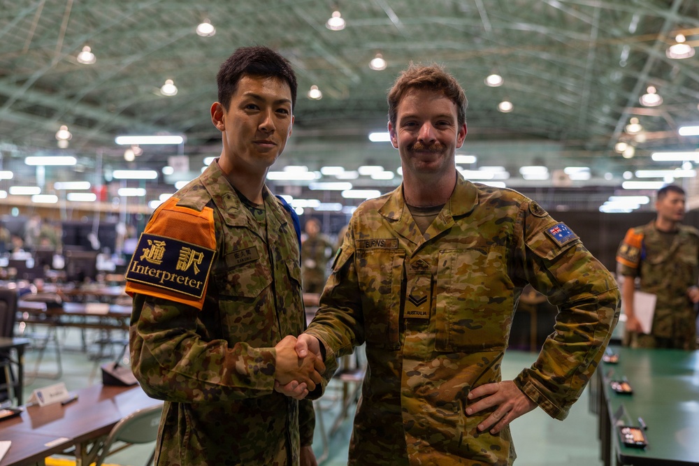 Two military personnel from different nations are shaking hands and posing for a photo inside a large military operations facility. The soldier on the left wears a Japanese Self-Defense Force uniform with an orange 'Interpreter' armband (通訳), while the soldier on the right wears an Australian Army uniform with the Australian flag patch and a name tape reading 'BURNS'. The setting appears to be a joint military exercise or multinational operations center.