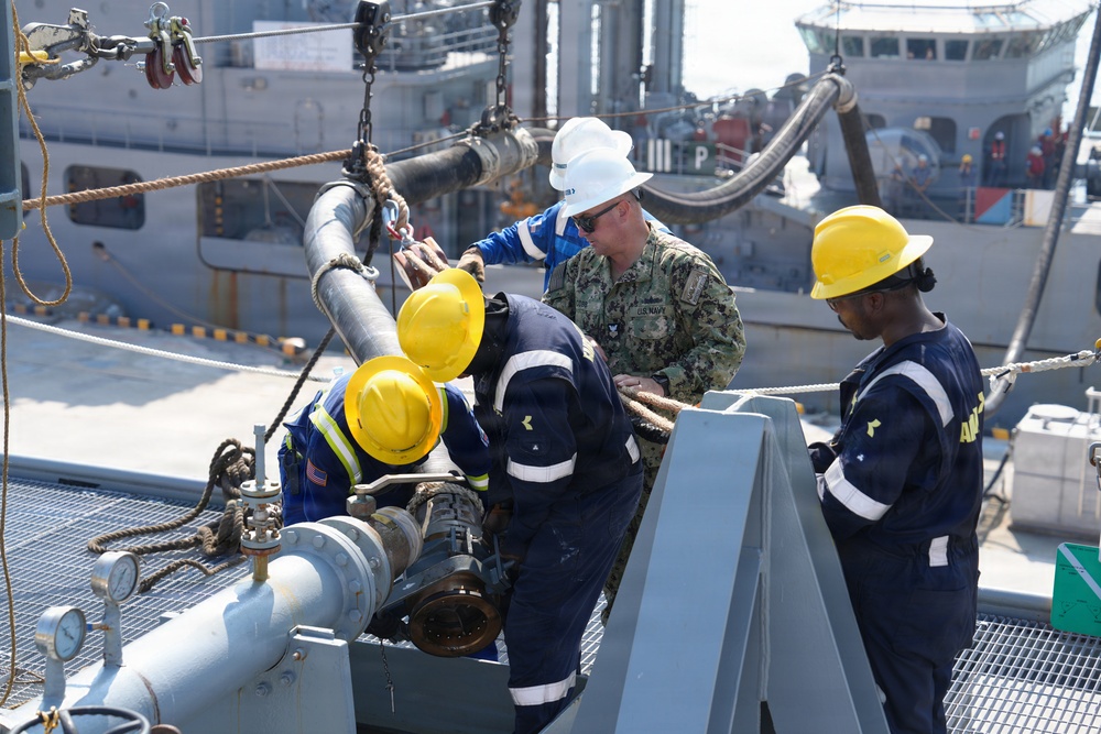 A U.S. Navy sailor in digital camouflage uniform and white hard hat is working alongside several personnel wearing yellow hard hats and dark blue coveralls on the deck of a naval vessel. They appear to be conducting a replenishment-at-sea (RAS) or fueling operation, connecting or disconnecting a large fuel transfer hose to deck fittings. A second vessel is visible in the background, consistent with an underway replenishment or pier-side fueling evolution.