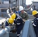A U.S. Navy sailor in digital camouflage uniform and white hard hat is working alongside several personnel wearing yellow hard hats and dark blue coveralls on the deck of a naval vessel. They appear to be conducting a replenishment-at-sea (RAS) or fueling operation, connecting or disconnecting a large fuel transfer hose to deck fittings. A second vessel is visible in the background, consistent with an underway replenishment or pier-side fueling evolution.