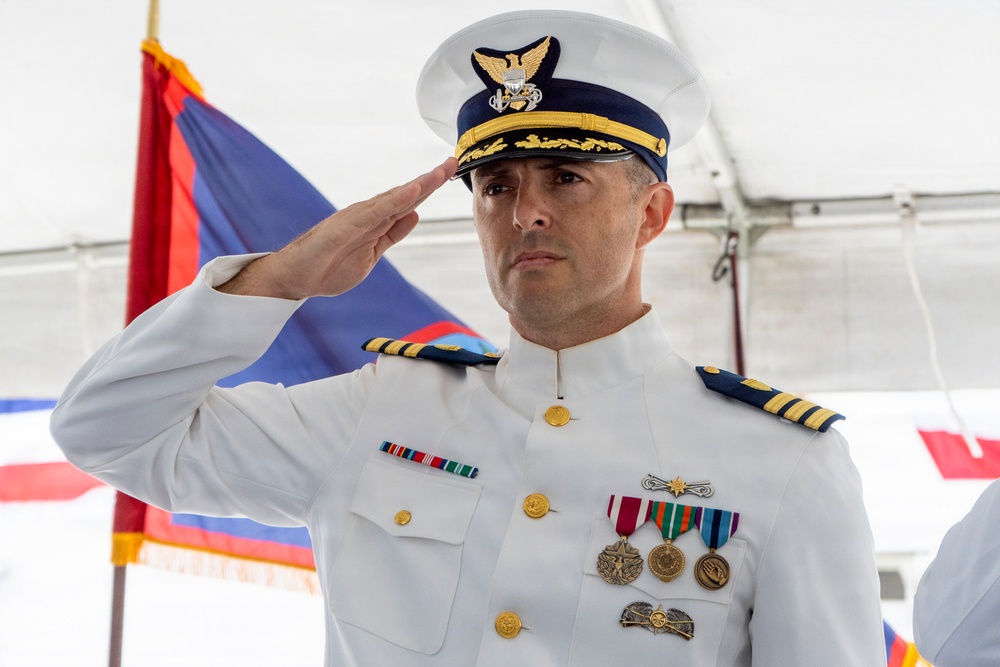A U.S. Coast Guard officer in dress white uniform is rendering a formal salute during what appears to be an official ceremony. The officer displays multiple service medals and ribbons, and wears rank insignia consistent with a senior officer grade. The setting appears to be an outdoor event under a tent with ceremonial flags visible in the background.