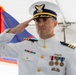 A U.S. Coast Guard officer in dress white uniform is rendering a formal salute during what appears to be an official ceremony. The officer displays multiple service medals and ribbons, and wears rank insignia consistent with a senior officer grade. The setting appears to be an outdoor event under a tent with ceremonial flags visible in the background.