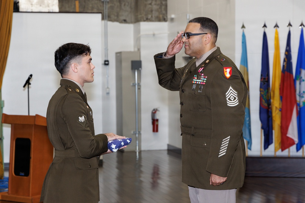 Two U.S. Army soldiers are participating in a formal military ceremony, likely a retirement or awards ceremony. The senior soldier on the right, wearing the rank of Sergeant Major (chevrons visible on sleeve) with multiple service ribbons and medals, is rendering a salute. The junior soldier on the left is holding a folded American flag, a common element in military retirement ceremonies.