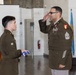Two U.S. Army soldiers are participating in a formal military ceremony, likely a retirement or awards ceremony. The senior soldier on the right, wearing the rank of Sergeant Major (chevrons visible on sleeve) with multiple service ribbons and medals, is rendering a salute. The junior soldier on the left is holding a folded American flag, a common element in military retirement ceremonies.