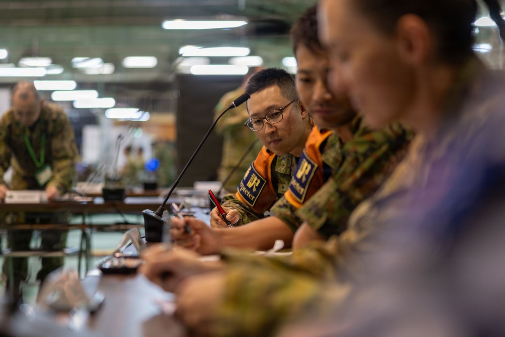 Military personnel are seated at a conference table during what appears to be a joint military briefing or planning session. At least two individuals are wearing orange vests with patches reading 'Interpreter' in both English and Japanese characters (通訳), indicating this is a multinational or bilateral military exercise or meeting. The setting appears to be a large indoor facility such as a command center or gymnasium.