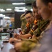 Military personnel are seated at a conference table during what appears to be a joint military briefing or planning session. At least two individuals are wearing orange vests with patches reading 'Interpreter' in both English and Japanese characters (通訳), indicating this is a multinational or bilateral military exercise or meeting. The setting appears to be a large indoor facility such as a command center or gymnasium.