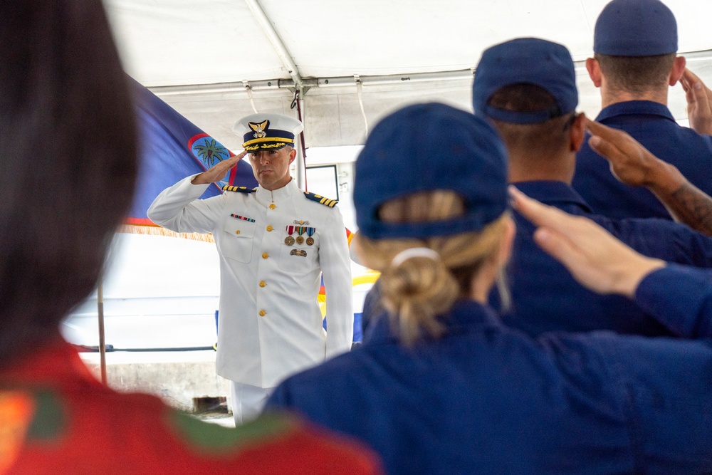 A military ceremony is taking place under a white tent, where a senior officer in a white dress uniform is rendering a salute to a formation of personnel in blue uniforms and caps. The officer displays multiple medals and rank insignia consistent with a Coast Guard commander or captain. A flag that appears to be the flag of Guam is visible in the background.