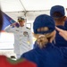 A military ceremony is taking place under a white tent, where a senior officer in a white dress uniform is rendering a salute to a formation of personnel in blue uniforms and caps. The officer displays multiple medals and rank insignia consistent with a Coast Guard commander or captain. A flag that appears to be the flag of Guam is visible in the background.