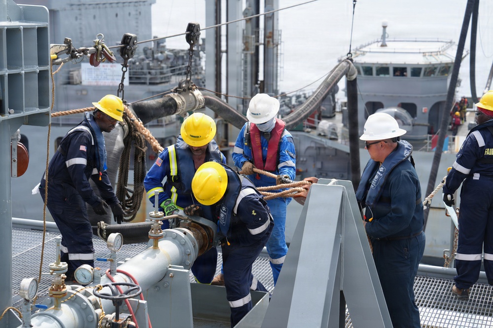 A crew of maritime workers is conducting a replenishment-at-sea (RAS) or fueling operation on the deck of a naval or auxiliary vessel. Multiple personnel wearing hard hats and safety gear are handling large hoses, ropes, and fuel transfer equipment between two ships positioned alongside each other. The operation appears to be an underway replenishment or dockside fuel transfer exercise.