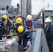 A crew of maritime workers is conducting a replenishment-at-sea (RAS) or fueling operation on the deck of a naval or auxiliary vessel. Multiple personnel wearing hard hats and safety gear are handling large hoses, ropes, and fuel transfer equipment between two ships positioned alongside each other. The operation appears to be an underway replenishment or dockside fuel transfer exercise.