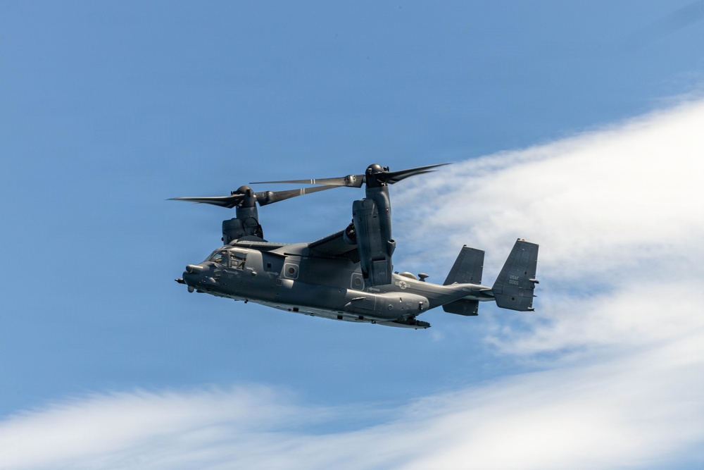 A military tiltrotor aircraft, identifiable as a Bell Boeing V-22 Osprey, is captured in flight against a partly cloudy blue sky. The aircraft is painted in a dark gray/charcoal color scheme consistent with U.S. military special operations use. The rotors are in a transitional or forward-flight position, and the image is sharp and well-exposed.
