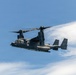 A military tiltrotor aircraft, identifiable as a Bell Boeing V-22 Osprey, is captured in flight against a partly cloudy blue sky. The aircraft is painted in a dark gray/charcoal color scheme consistent with U.S. military special operations use. The rotors are in a transitional or forward-flight position, and the image is sharp and well-exposed.