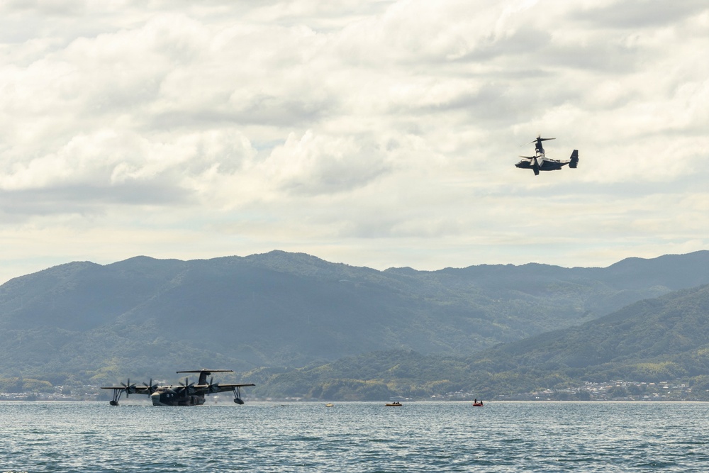 The image shows two distinct military aircraft over a calm body of water with green mountainous terrain in the background. A large flying boat/seaplane is resting on the water surface, while a tiltrotor aircraft (consistent with a V-22 Osprey type) is airborne in the upper right. Several small watercraft are also visible on the water near the seaplane.