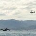 The image shows two distinct military aircraft over a calm body of water with green mountainous terrain in the background. A large flying boat/seaplane is resting on the water surface, while a tiltrotor aircraft (consistent with a V-22 Osprey type) is airborne in the upper right. Several small watercraft are also visible on the water near the seaplane.