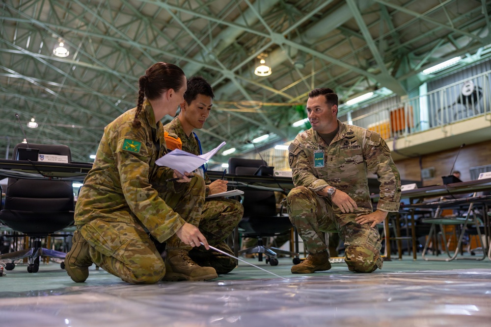 Three military personnel from different nations are crouched on a floor in what appears to be a large indoor facility or hangar, engaged in a planning or briefing session. Two individuals wearing Australian Army camouflage uniforms and one wearing US Army MultiCam uniform are examining documents and what appears to be a map or diagram on the floor. This appears to be a multinational military exercise or joint planning event.