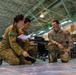 Three military personnel from different nations are crouched on a floor in what appears to be a large indoor facility or hangar, engaged in a planning or briefing session. Two individuals wearing Australian Army camouflage uniforms and one wearing US Army MultiCam uniform are examining documents and what appears to be a map or diagram on the floor. This appears to be a multinational military exercise or joint planning event.