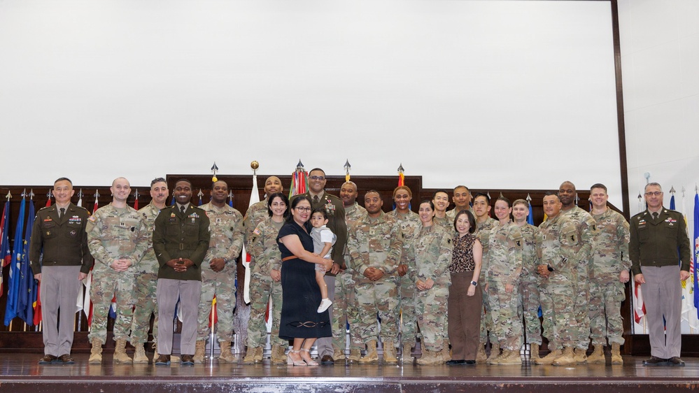 A formal military group photograph taken indoors, likely during a ceremony or official event. The group consists of approximately 20-25 individuals, predominantly in U.S. Army uniforms (OCP/multicam pattern), with two civilians including a woman holding a young child positioned at the center. Multiple national and military flags are displayed on a stage backdrop behind the group.