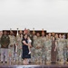A formal military group photograph taken indoors, likely during a ceremony or official event. The group consists of approximately 20-25 individuals, predominantly in U.S. Army uniforms (OCP/multicam pattern), with two civilians including a woman holding a young child positioned at the center. Multiple national and military flags are displayed on a stage backdrop behind the group.