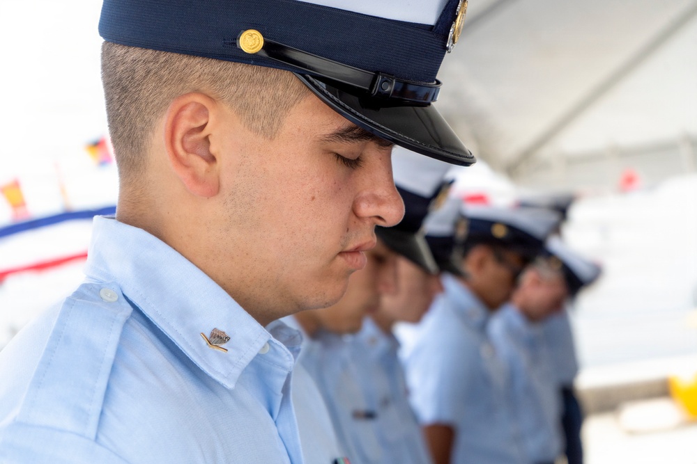 A row of uniformed personnel, likely U.S. Coast Guard members, stand with heads bowed in what appears to be a formal ceremony or moment of silence. The foreground subject is a young male in a light blue uniform shirt and dark navy peaked cap with gold insignia. Multiple personnel are visible in the background, slightly out of focus, wearing identical uniforms.