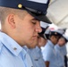 A row of uniformed personnel, likely U.S. Coast Guard members, stand with heads bowed in what appears to be a formal ceremony or moment of silence. The foreground subject is a young male in a light blue uniform shirt and dark navy peaked cap with gold insignia. Multiple personnel are visible in the background, slightly out of focus, wearing identical uniforms.
