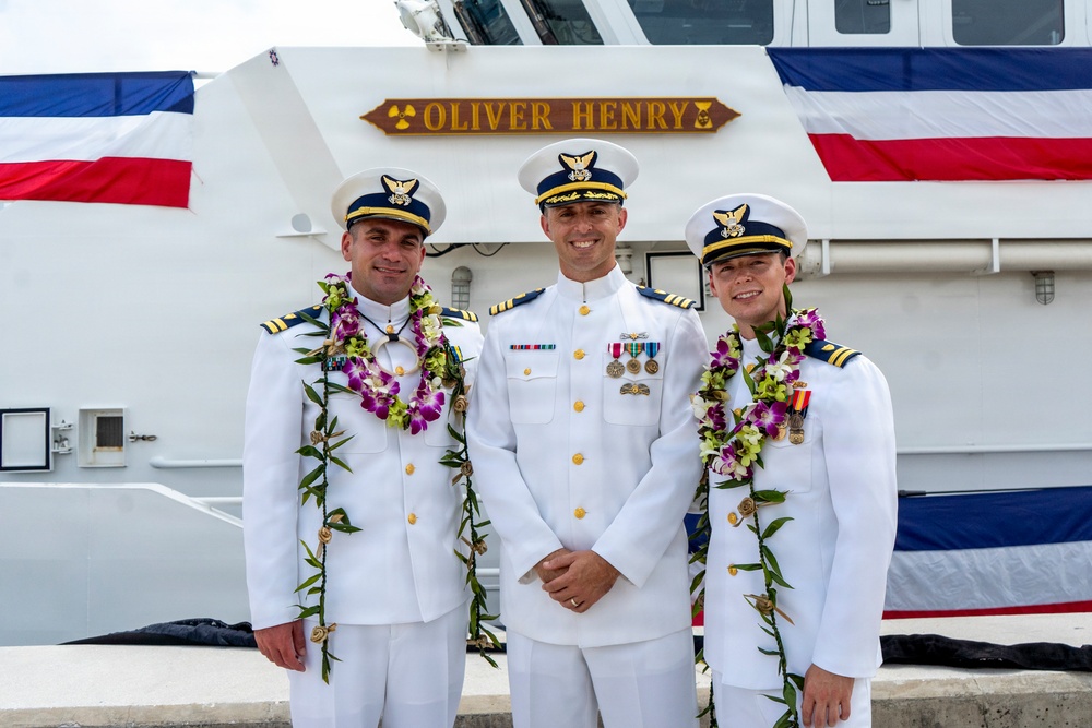 Three U.S. Coast Guard officers in white dress uniforms pose for a formal photograph in front of a vessel named 'Oliver Henry.' Two of the officers are wearing traditional Hawaiian lei garlands, suggesting a commissioning or change-of-command ceremony. The setting appears to be a dock or pier, with the ship's nameplate and a red, white, and blue flag visible in the background.
