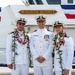 Three U.S. Coast Guard officers in white dress uniforms pose for a formal photograph in front of a vessel named 'Oliver Henry.' Two of the officers are wearing traditional Hawaiian lei garlands, suggesting a commissioning or change-of-command ceremony. The setting appears to be a dock or pier, with the ship's nameplate and a red, white, and blue flag visible in the background.