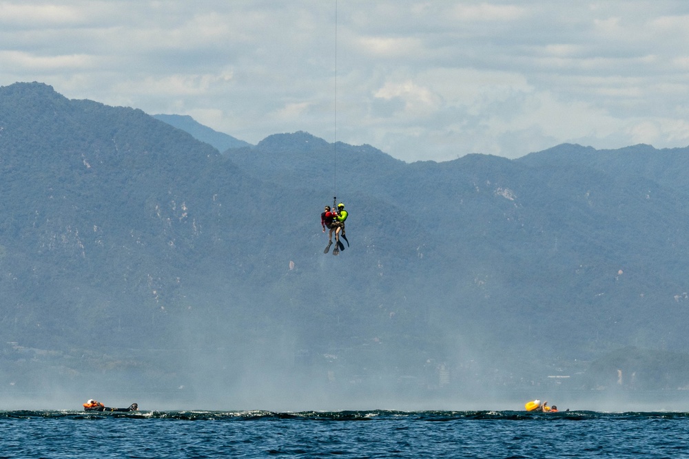 Two individuals are suspended mid-air over a body of water, appearing to be involved in a helicopter hoist or rescue operation. One person is wearing a bright yellow-green vest and the other a red jacket, connected by a cable extending upward out of frame. Below on the water surface, two small watercraft or inflatable vessels are visible on either side.