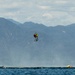 Two individuals are suspended mid-air over a body of water, appearing to be involved in a helicopter hoist or rescue operation. One person is wearing a bright yellow-green vest and the other a red jacket, connected by a cable extending upward out of frame. Below on the water surface, two small watercraft or inflatable vessels are visible on either side.